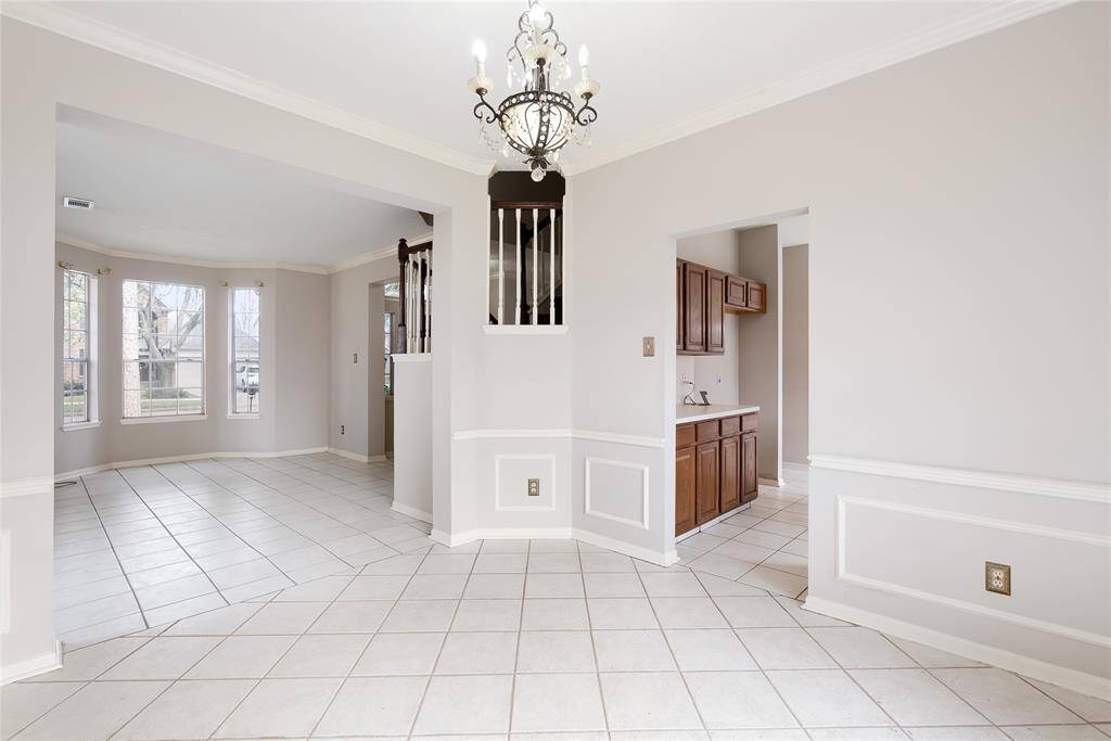 This photo showcases a bright and open living area with light-colored tile flooring, neutral walls, and decorative molding. It features a central chandelier, ample natural light from large windows, and easy access to an adjacent room with wooden cabinetry.