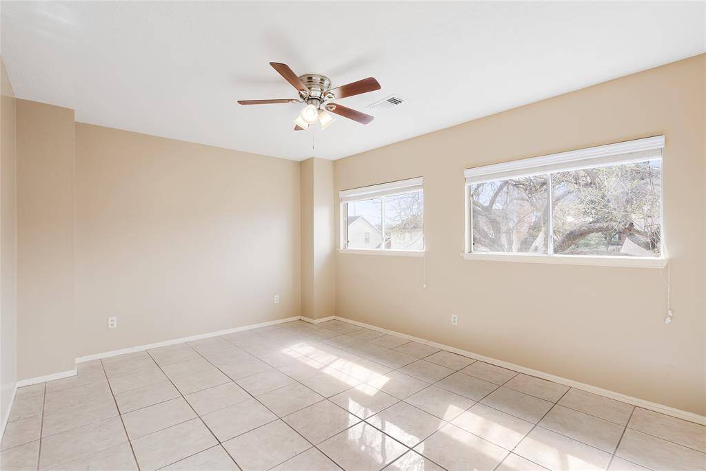 This room is bright and airy, featuring neutral walls and a tiled floor. It has ample natural light from two large windows and includes a ceiling fan for added comfort.