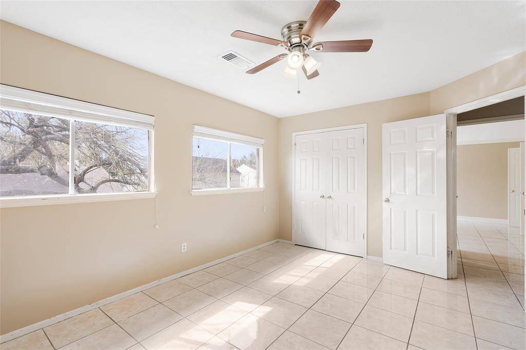 Bright room with cream walls, tiled floor, and a ceiling fan. The space features two windows for natural light and a closet with double doors, creating a clean and airy feel.