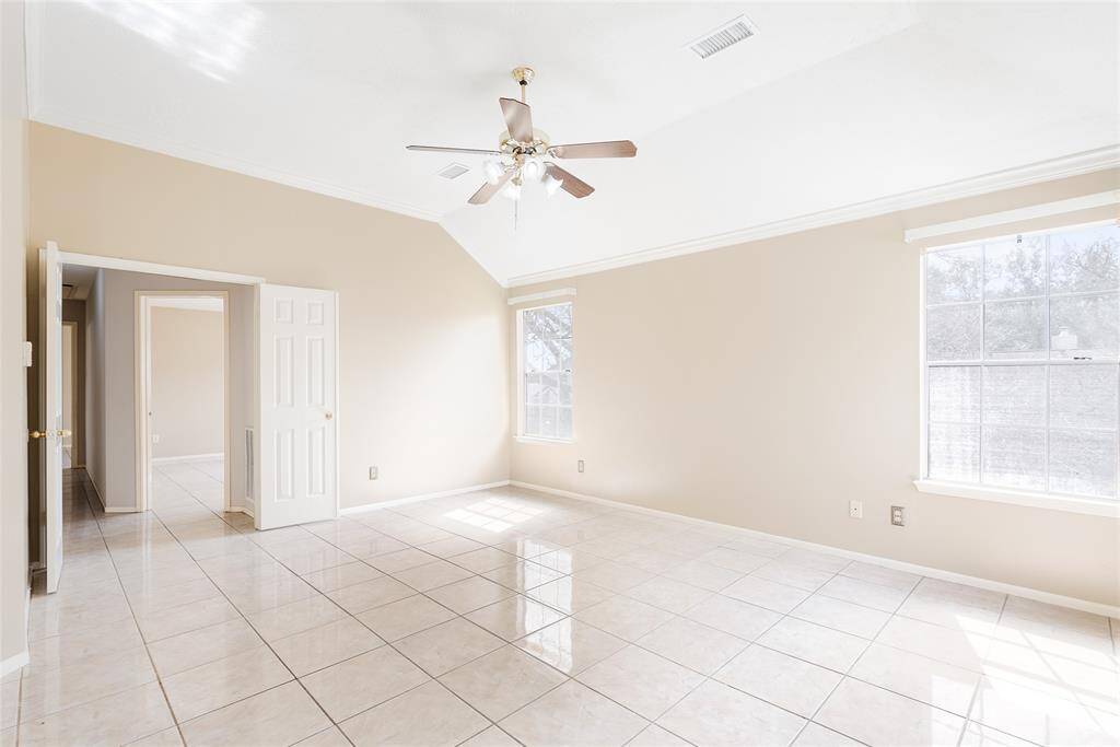 Bright and spacious room with a vaulted ceiling and a ceiling fan. It features large windows that let in natural light, neutral walls, and shiny tiled flooring. The space connects to adjacent rooms through open doorways, creating a seamless flow.