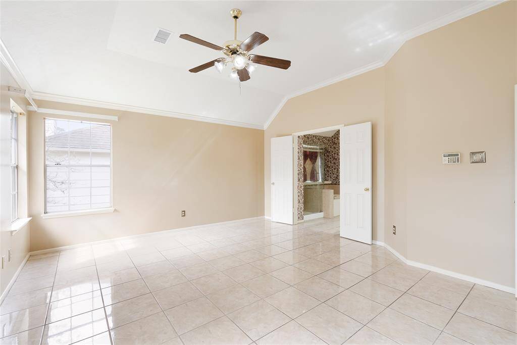 This spacious room features neutral walls, tiled flooring, and a ceiling fan. Natural light streams through a large window, enhancing the open feel. Double doors lead to a bathroom with elegant wallpaper and a bathtub.