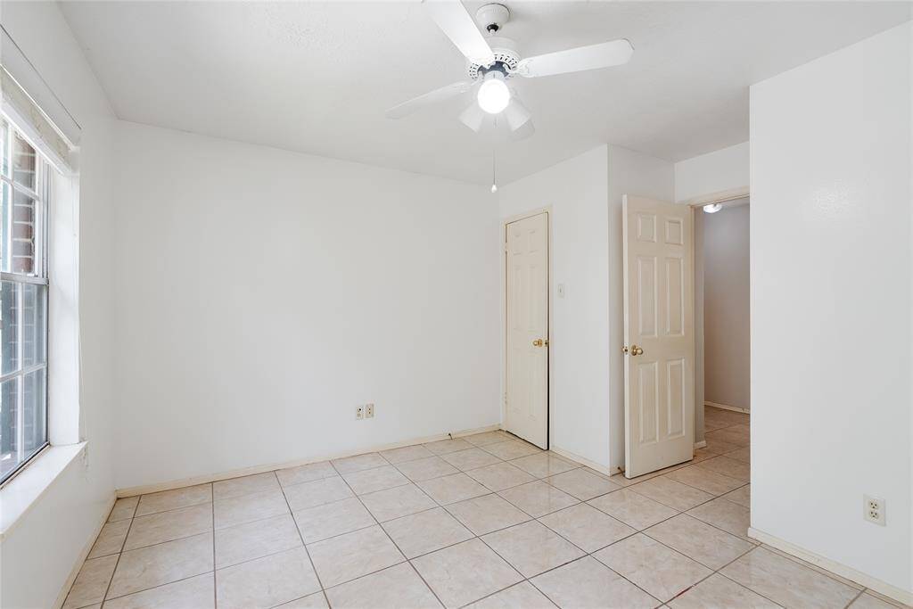 This room is bright and airy with white walls and tiled flooring. It features a ceiling fan and a large window for natural light. There are two doors, leading to a closet and hallway.