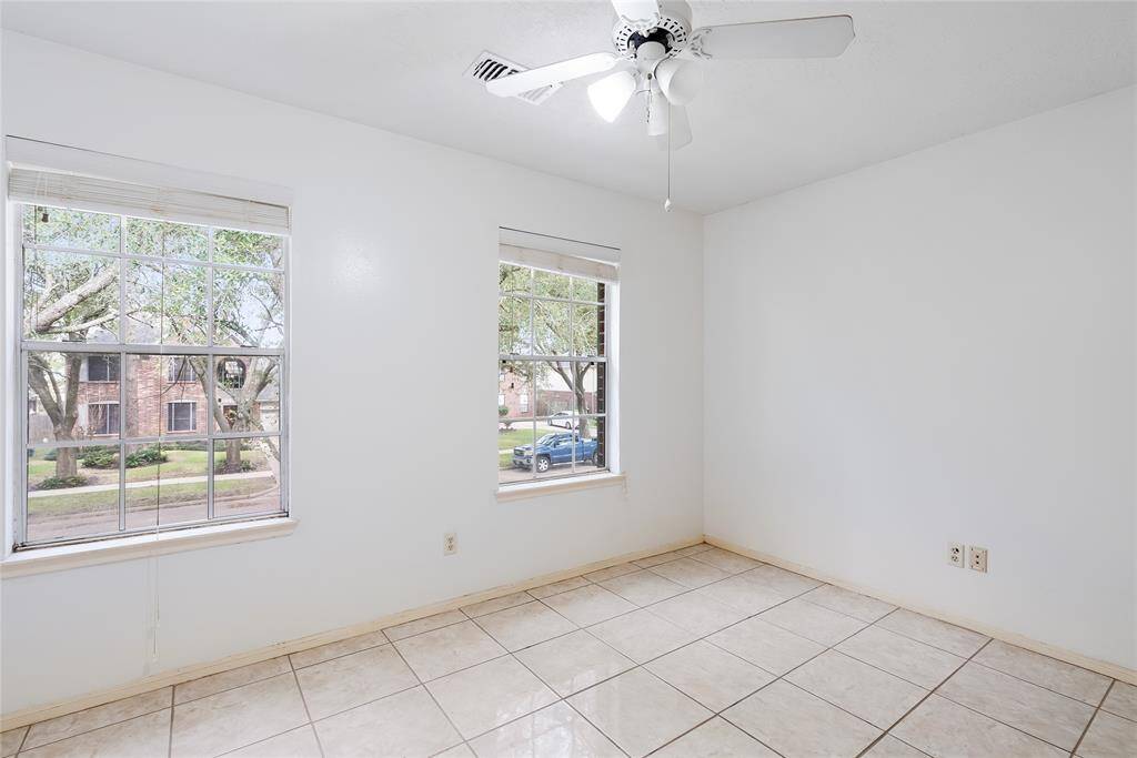 This room is bright and airy, featuring neutral walls and a tiled floor. It has ample natural light from two large windows and includes a ceiling fan for added comfort.