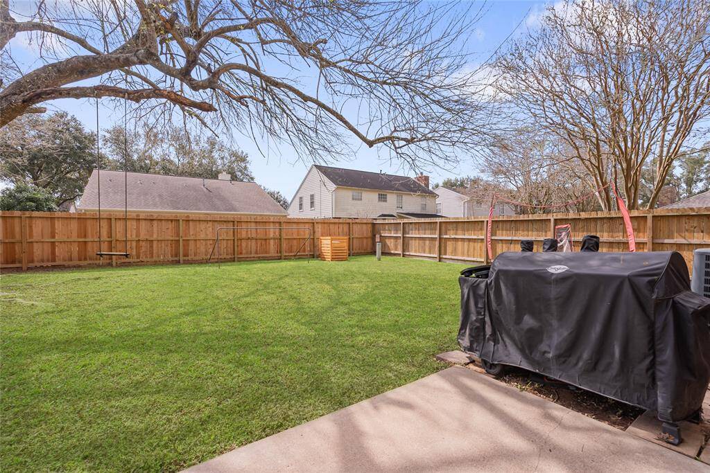 Spacious backyard with lush green lawn, surrounded by a wooden fence offering privacy. Features a large tree, a swing set, and a covered grill on a concrete patio area, perfect for outdoor activities and entertaining.