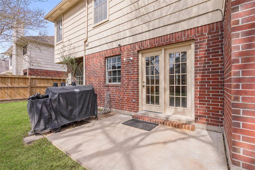 This photo showcases a cozy backyard patio with a brick exterior, featuring French doors leading into the house. The space includes a covered grill area and is bordered by a wooden privacy fence, offering a charming outdoor setting.