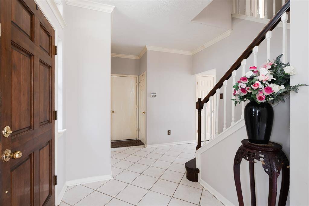 This inviting entryway features a wooden door, light gray walls, and white tile flooring. A staircase with a dark wood handrail and white balusters adds elegance, accompanied by a floral arrangement on a decorative stand, creating a welcoming atmosphere.