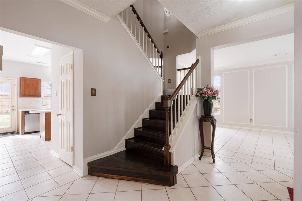 This photo showcases a bright, open foyer with a dark wood staircase. The space features light-colored tile flooring and neutral walls, leading into a kitchen area with visible wooden cabinets. A decorative stand with flowers adds charm to the entrance.