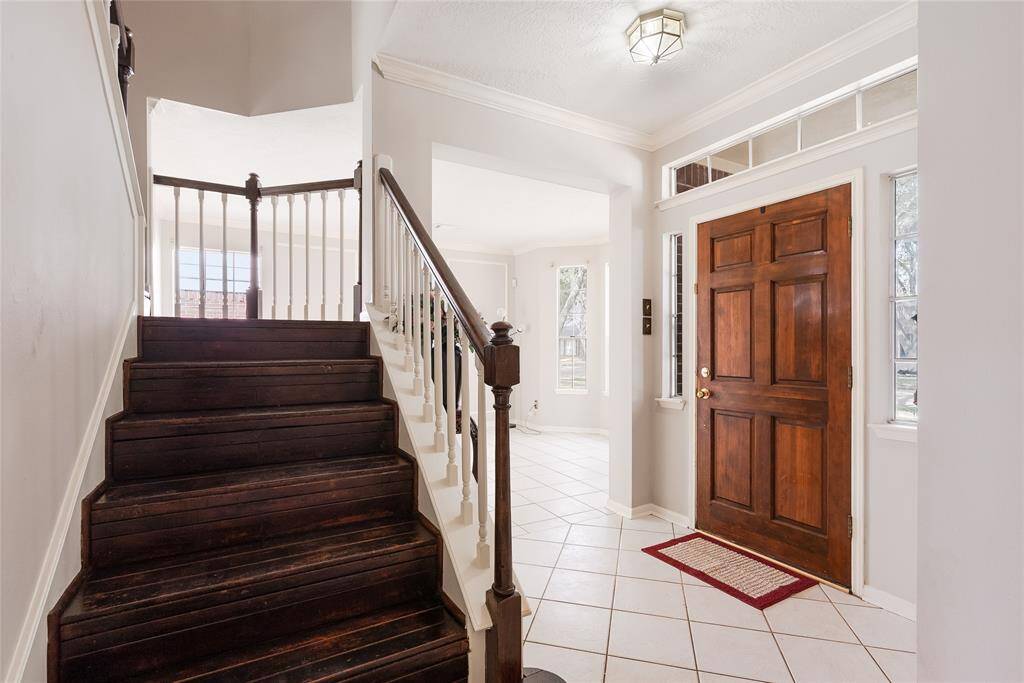 This photo showcases a welcoming entryway with a wooden front door and tiled flooring. The space is bright, featuring a staircase with dark wood steps and white railings, leading to an open living area with large windows.