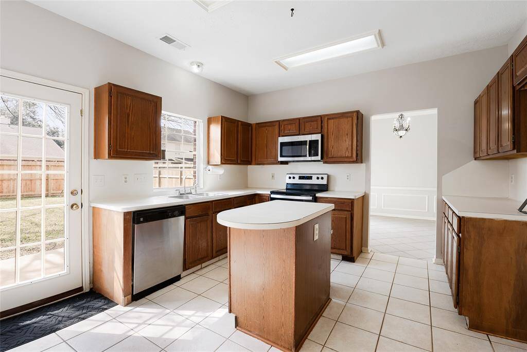 This kitchen features clean light countertops, wooden cabinets, and modern stainless steel appliances, including a dishwasher and stove. A central island offers additional workspace, while a door leads to the backyard, providing natural light and convenient outdoor access.
