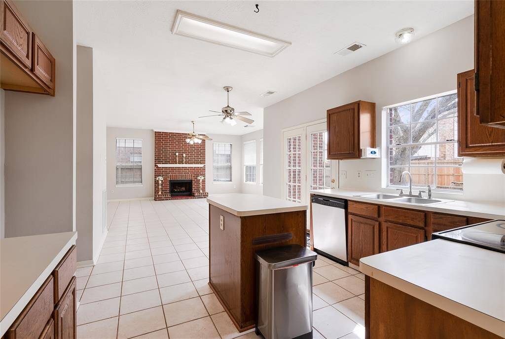 This bright, open-concept kitchen and living area features tiled floors, wooden cabinets, and a cozy brick fireplace. Large windows and glass doors provide plenty of natural light, creating a warm and inviting atmosphere.
