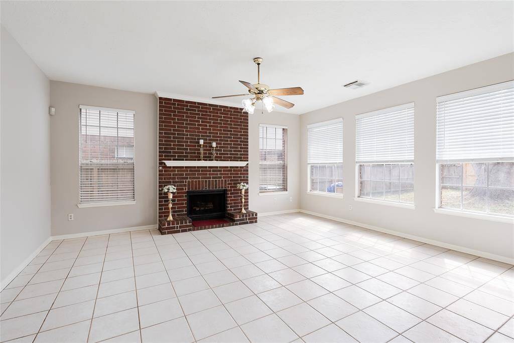 Bright and spacious living room featuring a brick fireplace, ceiling fan, and large windows for natural light. The room has a clean, neutral tile floor, creating a versatile space for any decor style.