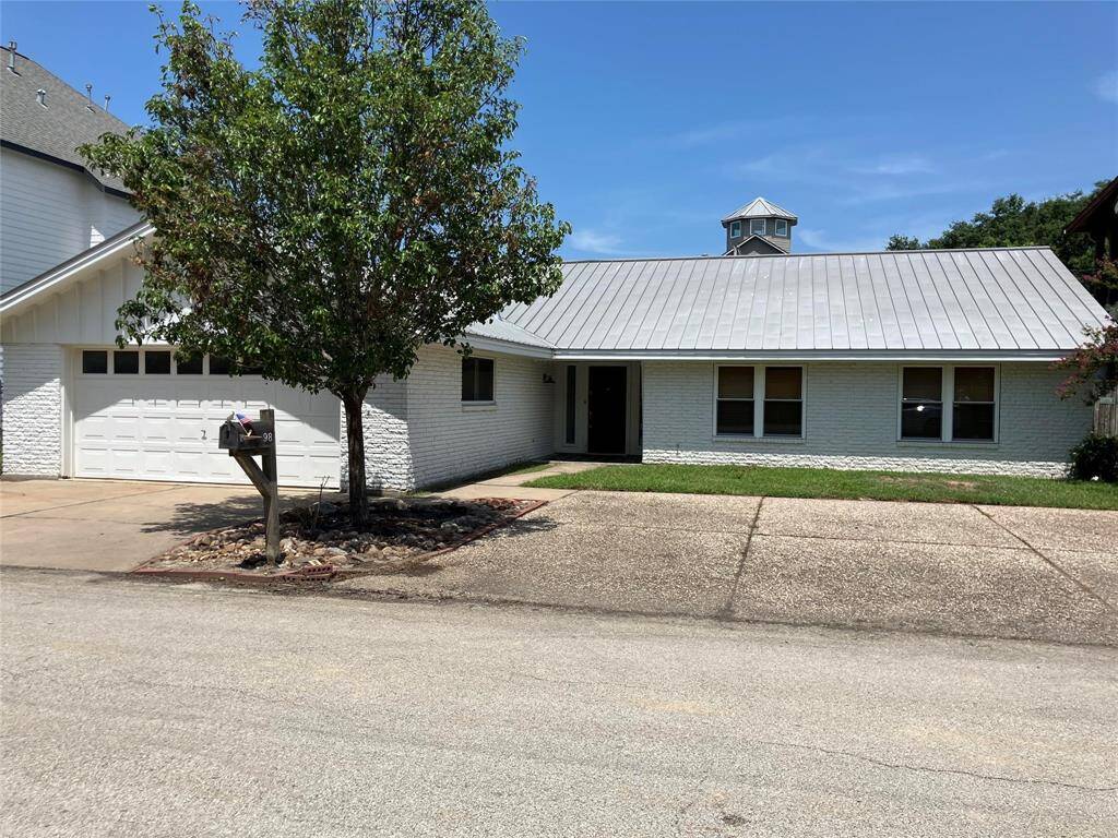 This photo shows a single-story home with a white brick exterior and a metal roof. It features a two-car garage, a small front lawn, and a tree by the driveway. The house has a clean, modern look and is situated on a quiet street.