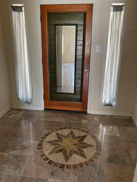 This entryway features a striking door with a wooden frame and glass panel, flanked by narrow windows with curtains. The floor is adorned with elegant marble tiles and a decorative medallion, creating a welcoming and sophisticated atmosphere.