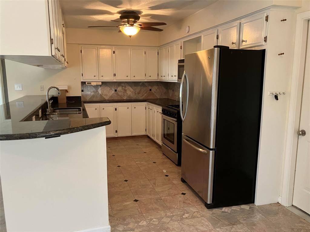 This kitchen features white cabinetry, a stainless steel refrigerator and oven, and a dark granite countertop. The space includes a ceiling fan, tile flooring, and a tiled backsplash, creating a modern and functional cooking area.