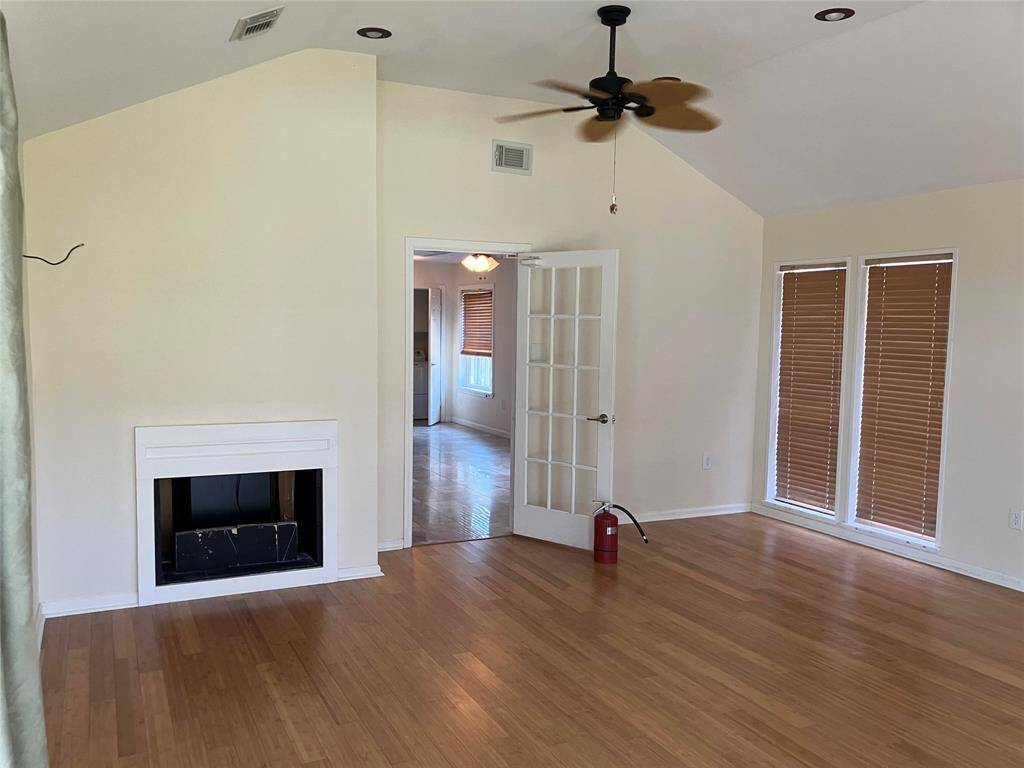 This room features a spacious area with wooden flooring, a ceiling fan, and a fireplace. It has large windows with blinds for natural light and a glass-paned door leading to an adjoining room.