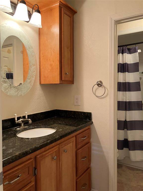 This bathroom features a dark granite countertop with a sink, wooden cabinets for storage, and a decorative mirror. It includes a well-lit vanity and a separate shower area with a striped curtain.
