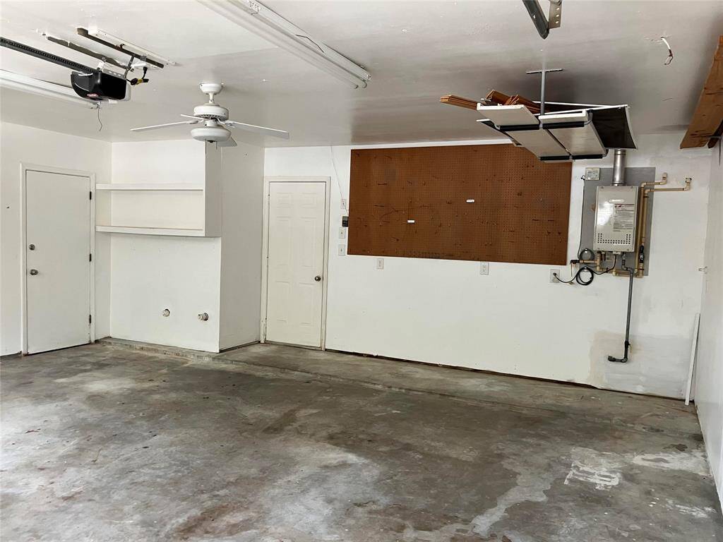 This is a spacious garage with a concrete floor, featuring a ceiling fan and overhead storage. It includes wall shelves, a pegboard for tools, and a tankless water heater. Ideal for parking, storage, or a workshop area.