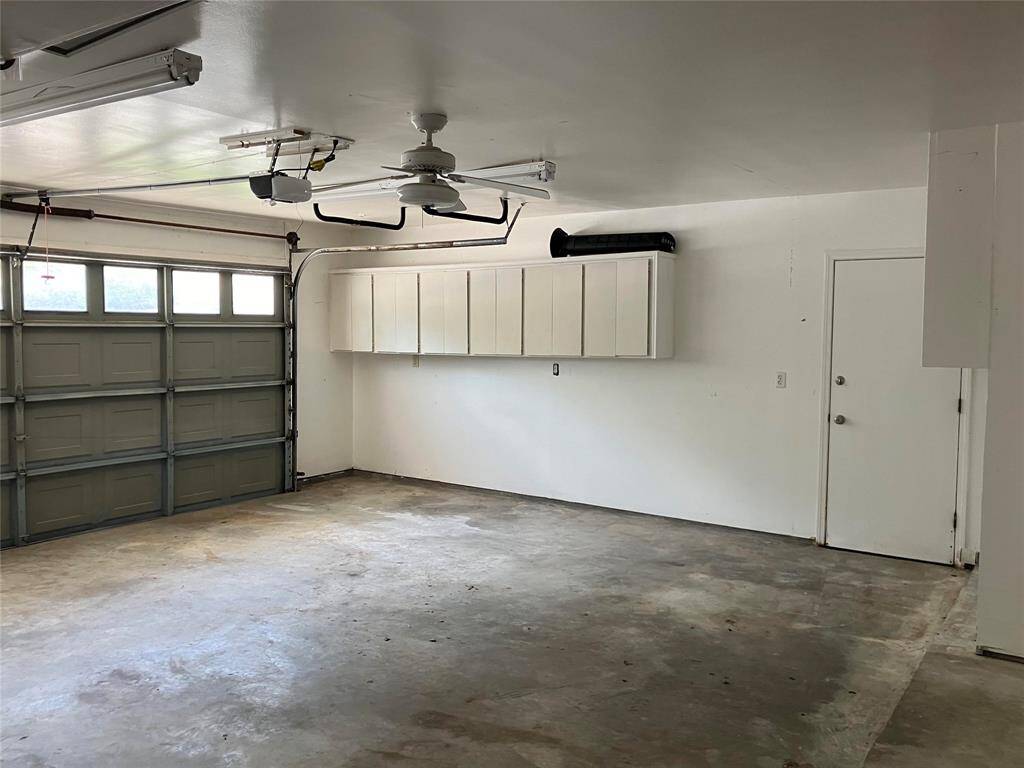 This photo shows a clean, spacious garage with a large overhead door, ceiling fan, and built-in storage cabinets. The floor is unfinished concrete, and there's a side door for additional access.