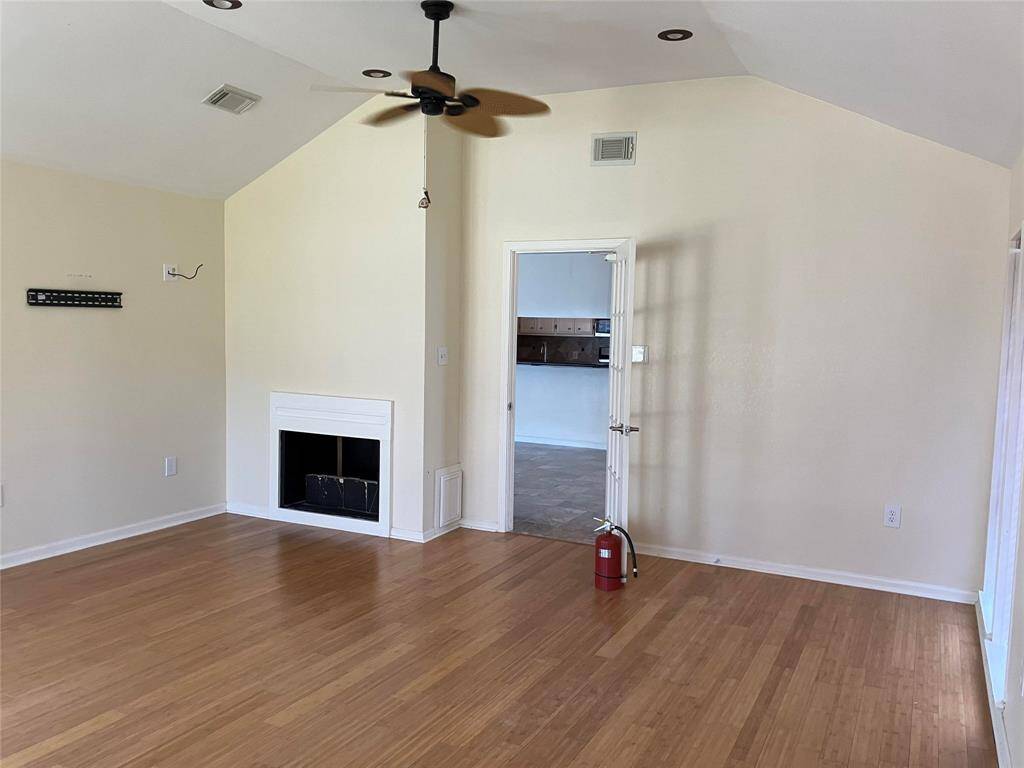 This spacious room features a vaulted ceiling with recessed lighting and a ceiling fan. It has a light hardwood floor and a small fireplace. A doorway leads to a tiled kitchen area, enhancing the open feel of the space.