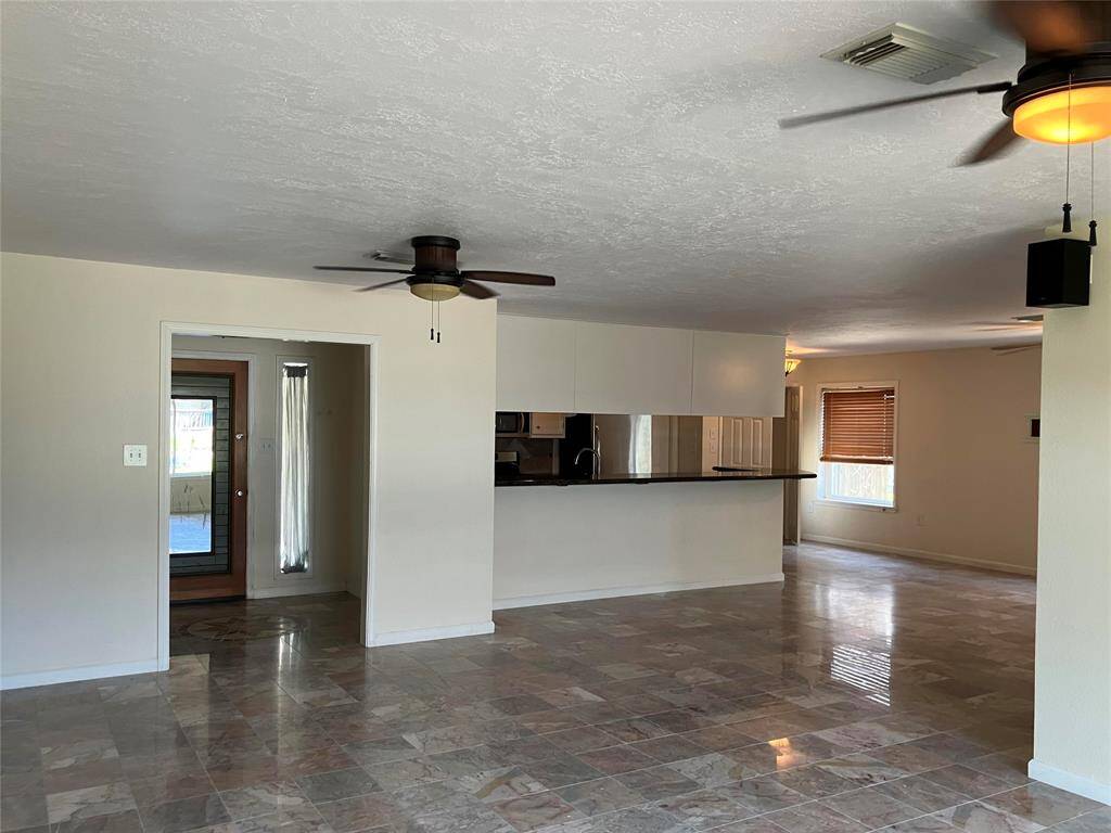 This spacious living area features a modern open layout with tiled floors, ceiling fans, and ample natural light. It includes a kitchen with a breakfast bar and a view of adjacent spaces, providing a welcoming and functional environment.
