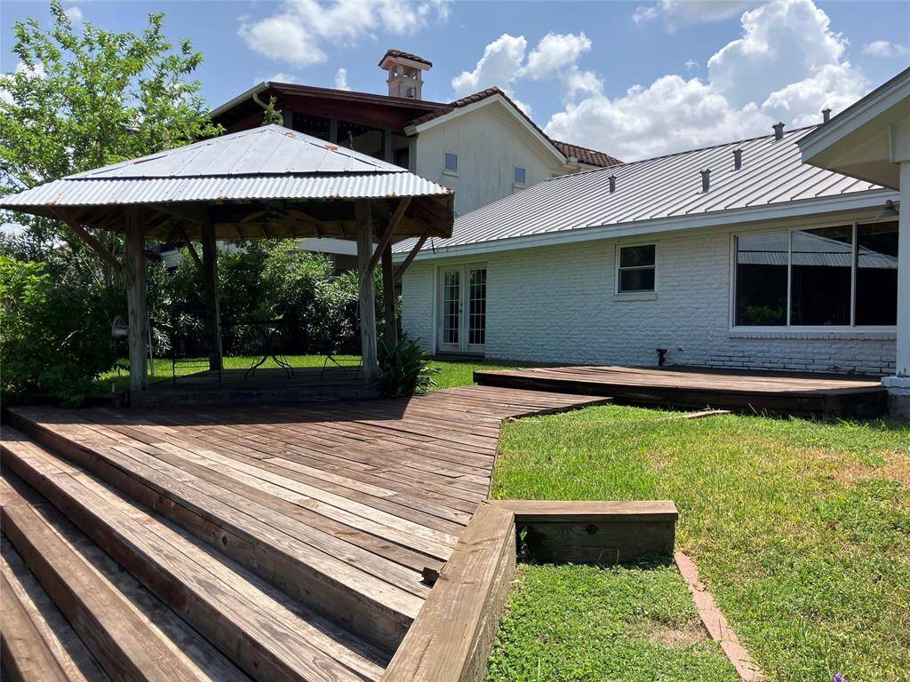 This photo showcases a backyard with a wooden deck and a gazebo, attached to a house with a white exterior and metal roof. The setting is sunny with a green lawn and some surrounding trees, providing a serene outdoor space.