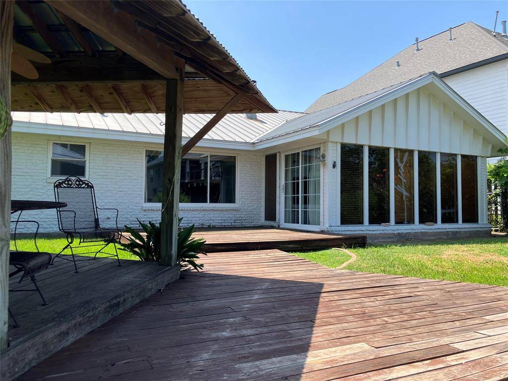 This photo showcases a charming backyard featuring a wooden deck and a covered patio area with seating. The exterior of the house is white brick with large windows, providing ample natural light. The yard has a grassy area bordered by plants, perfect for outdoor relaxation or entertaining.
