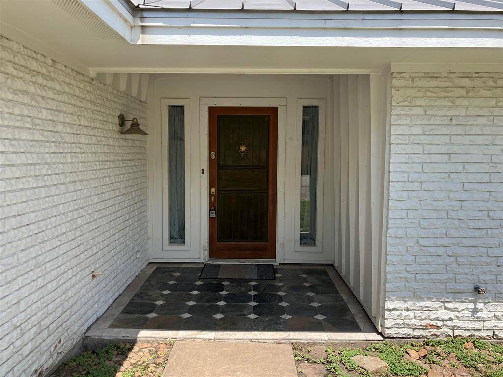 This image shows a welcoming front entrance with a wooden door and side windows. The area features a covered porch with patterned tile flooring and white brick walls, offering a clean and classic look. A metal wall-mounted light fixture adds character to the entryway.