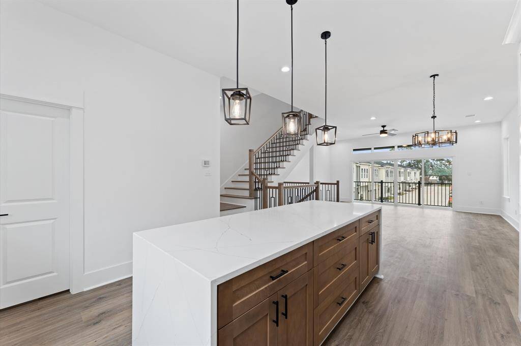 This modern kitchen features a spacious island with a sleek white countertop, pendant lighting, and a mix of white and wood cabinetry. The open layout is complemented by stainless steel appliances and light wood flooring, creating a bright and inviting space.