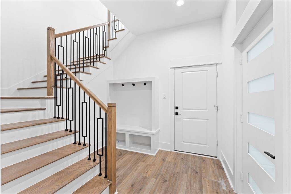 This photo showcases a modern, bright entryway featuring a wooden staircase with sleek black metal railings. The space has light wood flooring, a white door, and a built-in nook with storage and hooks, offering functionality and style.