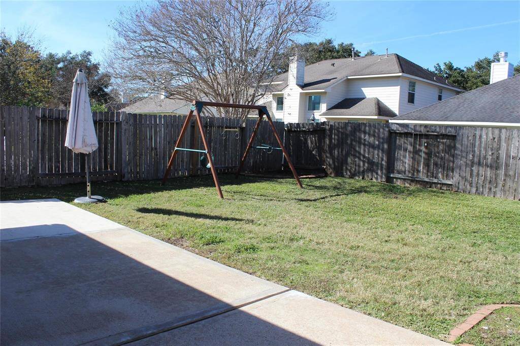 The photo shows a backyard with a concrete patio area, a grassy lawn, and a wooden swing set. The yard is enclosed by a wooden fence, offering privacy, and there's a large umbrella providing shade.