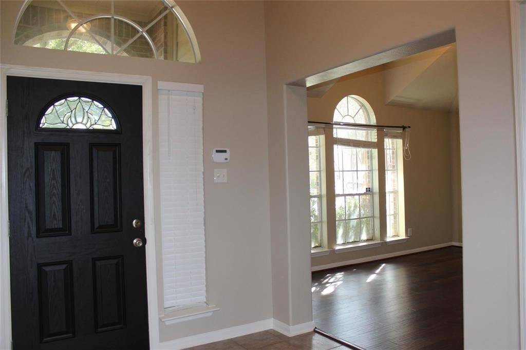 This photo shows a welcoming entryway with a stylish black door featuring decorative glass. Adjacent is a bright, open room with large arched windows, offering plenty of natural light and dark hardwood floors.