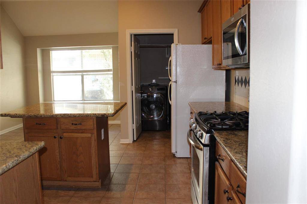 This kitchen features granite countertops, wooden cabinets, and a gas stove. It has a tile floor and an adjacent laundry area with a washer and dryer. A large window provides natural light.
