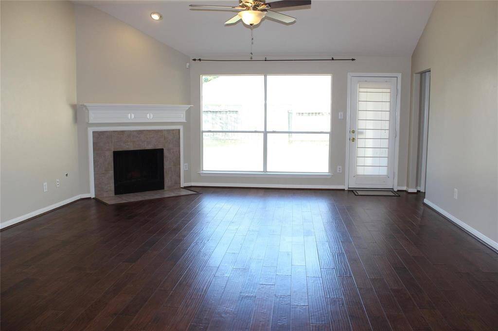 This living room features a high ceiling, dark hardwood floors, and a large window that allows plenty of natural light. It includes a fireplace with a white mantel and a ceiling fan for added comfort. The room also has a door leading outside, enhancing its functionality.