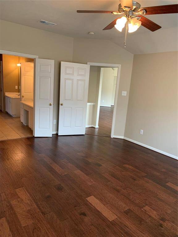 Another angle of Master bedroom, a spacious room with rich, dark wood flooring and neutral walls. It features a ceiling fan with light, a double-door entry to an adjoining room, and a door leading to a bathroom with visible vanity and tile flooring, creating a warm and inviting atmosphere.