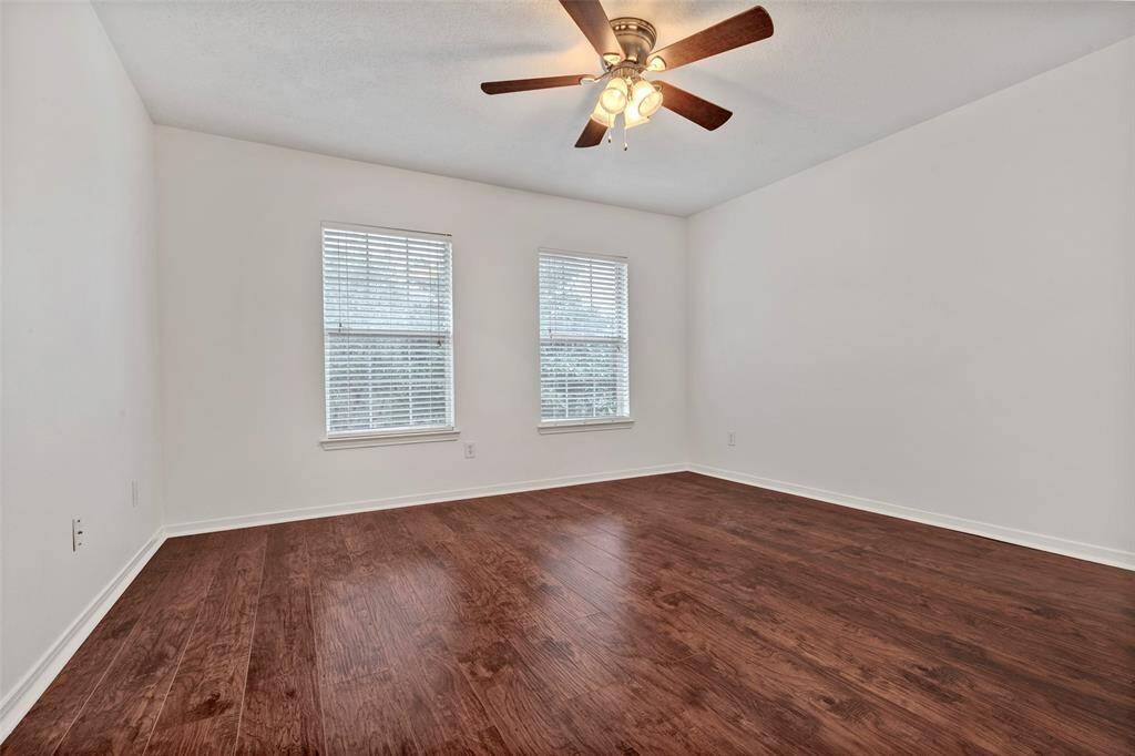 This room features clean white walls, rich wood flooring, and a ceiling fan with lights. Natural light fills the space through two windows with blinds, creating a bright and inviting atmosphere.