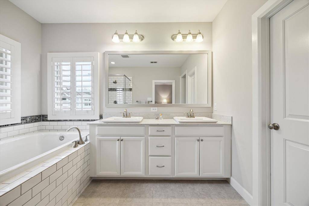 Bright and modern bathroom featuring a double vanity with white cabinetry, large mirror, and stylish light fixtures. It includes a spacious bathtub with elegant tile accents and a window with plantation shutters, creating a relaxing atmosphere.