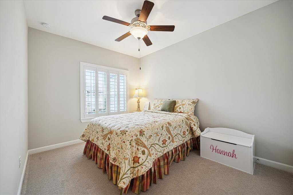 This cozy secondary bedroom features neutral walls, a ceiling fan, and a window with elegant plantation shutters that are throughout the home.