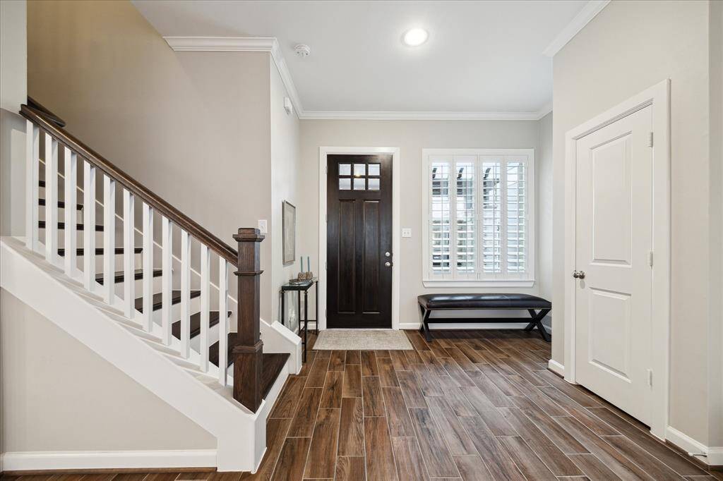 The entryway features wood-look tile flooring, a dark wooden front door, and a staircase with white railings. A window with shutters allows natural light. Neutral walls and a closet add to the functionality and style.