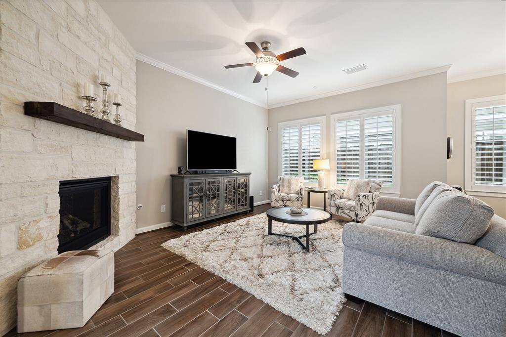 This cozy living room features a stone fireplace, wall of windows to the back yard, and a ceiling fan. Natural light filters through plantation shutters, enhancing the room's inviting atmosphere.