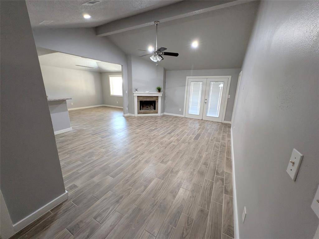 Spacious living area with wood-look tile flooring, a central fireplace, high ceilings, and French doors leading outside. The room is open and bright, offering a modern and inviting atmosphere.