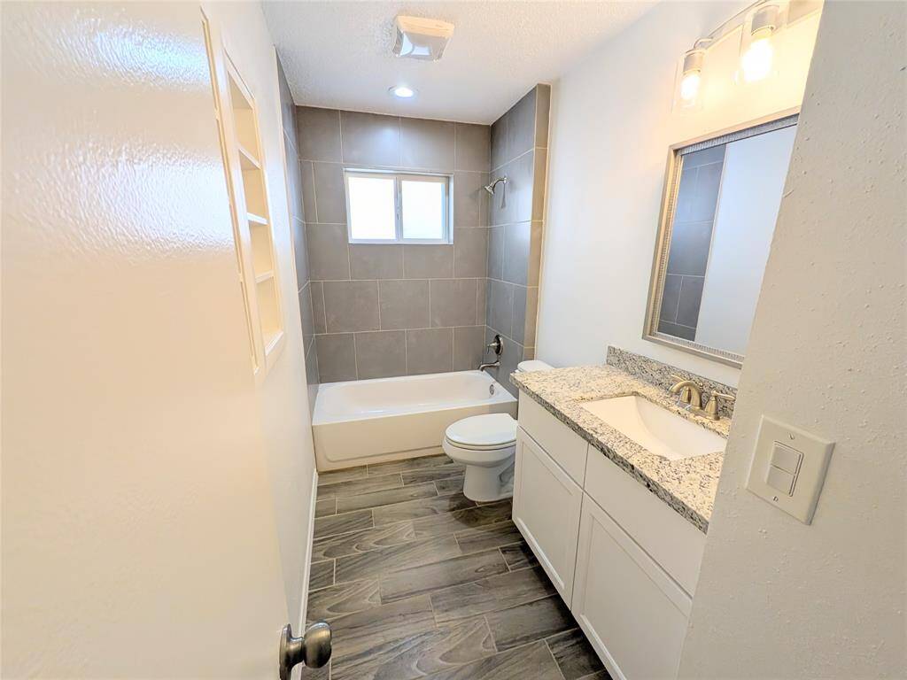 Seecondarry bathroom features a modern design with a sleek granite countertop, white vanity, and elegant lighting. The space includes a tiled shower-tub combo and wood-look flooring, providing a clean and contemporary feel.