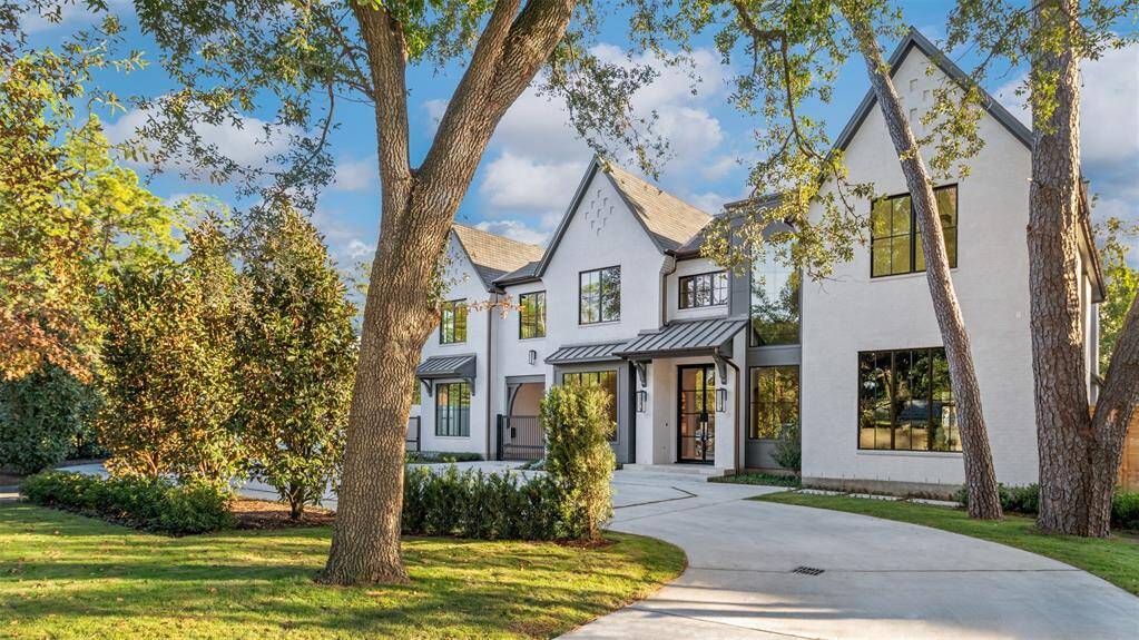 This is a modern, two-story home featuring a mix of white stucco and dark gray accents, large windows, and a striking gabled roofline. The front entrance has a welcoming feel with a covered porch area. The property includes a spacious driveway and a contemporary fence to the side, offering a blend of elegance and privacy.
