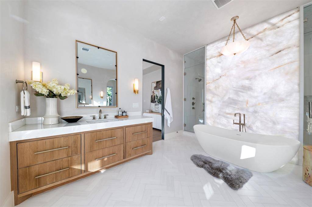 his luxury primary bathroom is a sanctuary of sophistication and indulgence. Custom fluted cabinets line the space, adding texture and timeless elegance to the design. The grand soaking tub is perfectly positioned beneath an onyx backlit wall, which softly illuminates the space with a warm, ambient glow, creating a spa-like atmosphere.