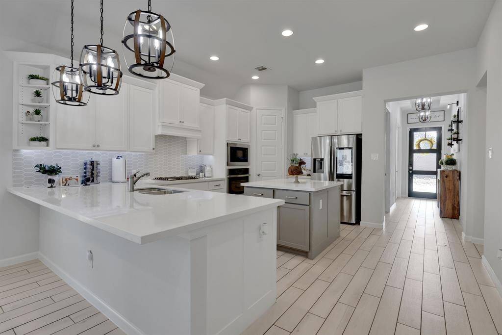 This modern kitchen features sleek white cabinetry, a large island with a sink, contemporary pendant lighting, and stainless steel appliances. The space is bright and open with a tiled floor, offering a clean and inviting atmosphere.