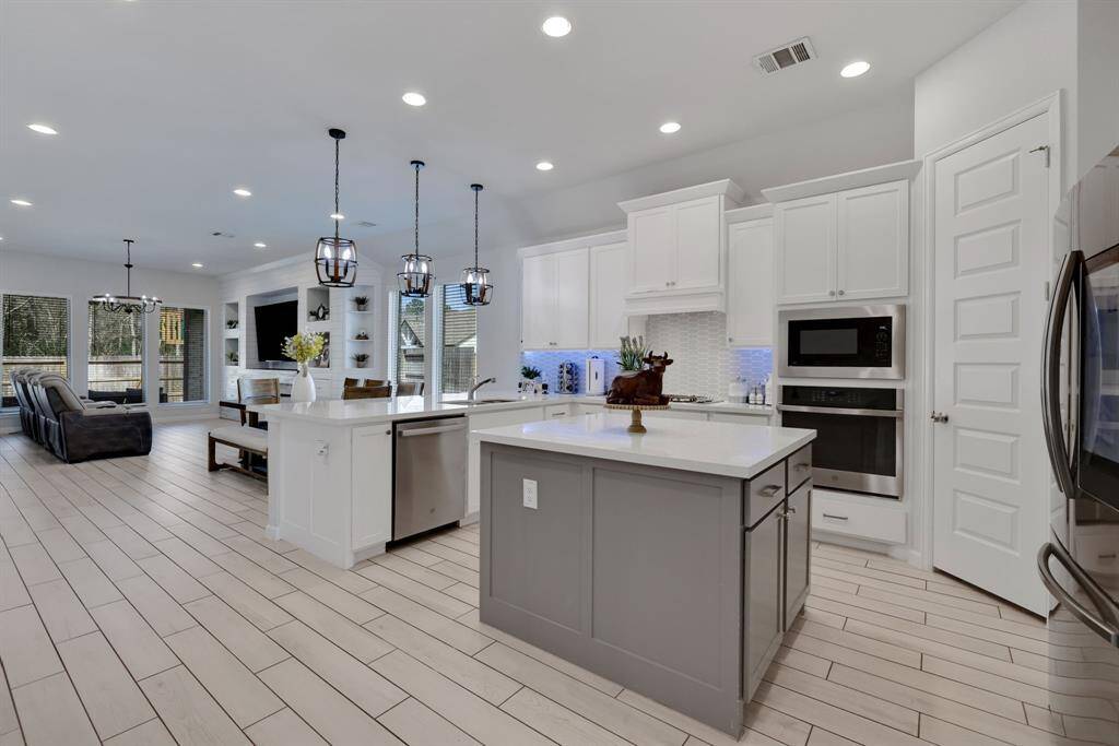 This open-concept kitchen and living area boasts modern design with white cabinetry, a spacious island, and stainless-steel appliances. The room is bright with ample recessed lighting and large windows, and it flows seamlessly into a cozy living space featuring built-in shelving and a decorative fireplace.