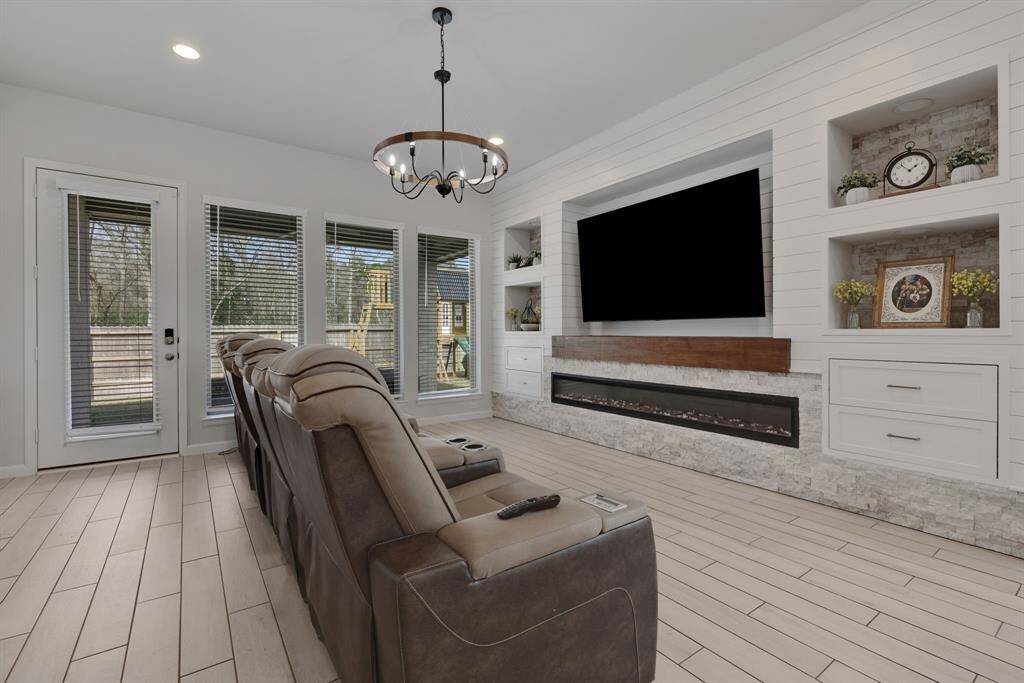 This spacious living room features a cozy, modern design with a large built-in entertainment center, an electric fireplace, and ample shelving for decor. The room is well-lit with natural light from the large windows and glass door leading to the backyard.