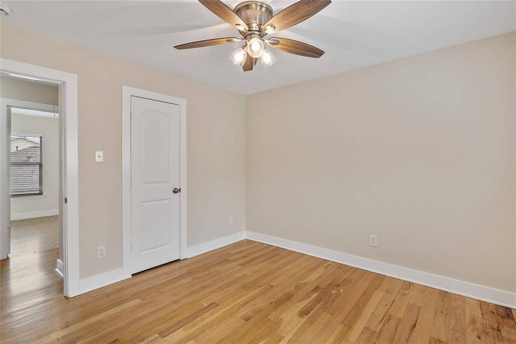 This room features hardwood floors, neutral walls, and a ceiling fan with lights. There's a white door and an open doorway leading to another room with a window, providing natural light. It's a cozy space suitable for various uses.