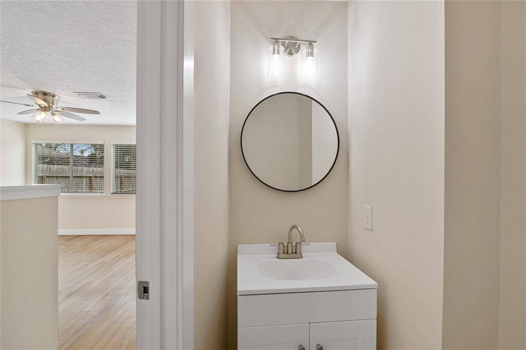 This photo shows a bright, cozy space with a small vanity area featuring a round mirror and modern light fixture. Adjacent is a living area with wood flooring, large windows for natural light, and a ceiling fan.