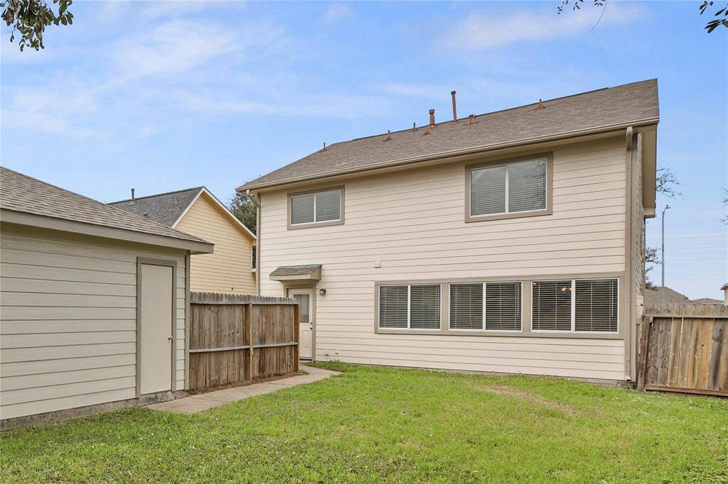 This photo shows the backyard of a two-story house with beige siding. It features a small grassy area, a wooden fence for privacy, and a detached structure on the left, possibly a shed or garage. The house has multiple windows, offering plenty of natural light.
