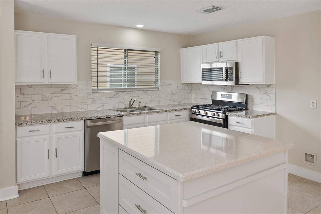 This kitchen features a modern design with white cabinetry, granite countertops, and a stylish marble backsplash. It includes stainless steel appliances, a gas range, and a central island, providing ample space for cooking and entertaining.